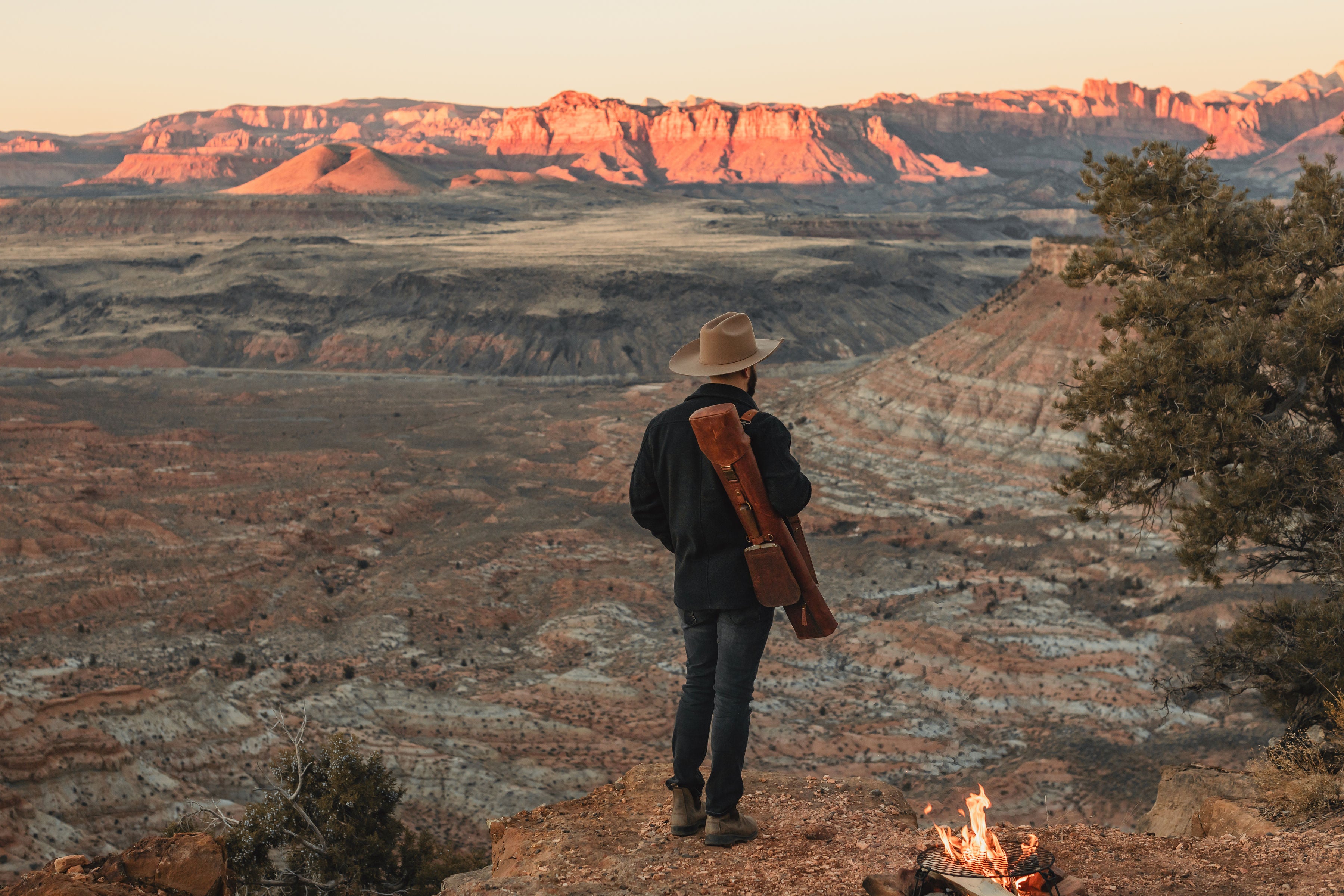 A man looking out at a mountain range with a Jack Sack on his back and a campfire behind him.
