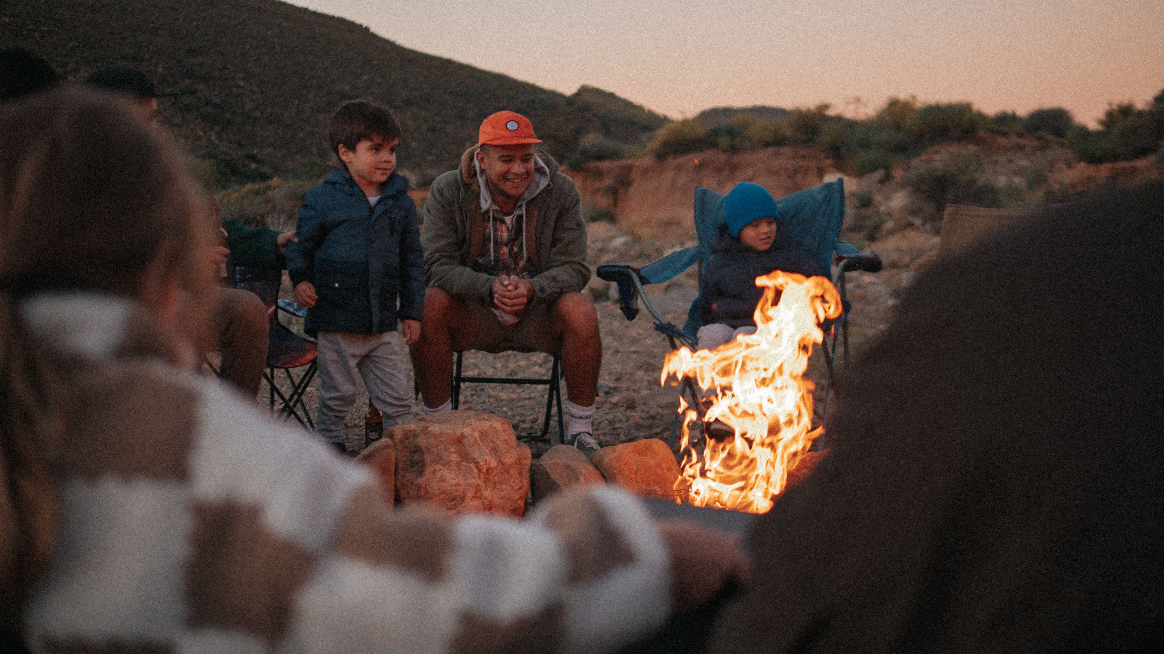 A dad sits in camping chairs with his two sons in front of a fire