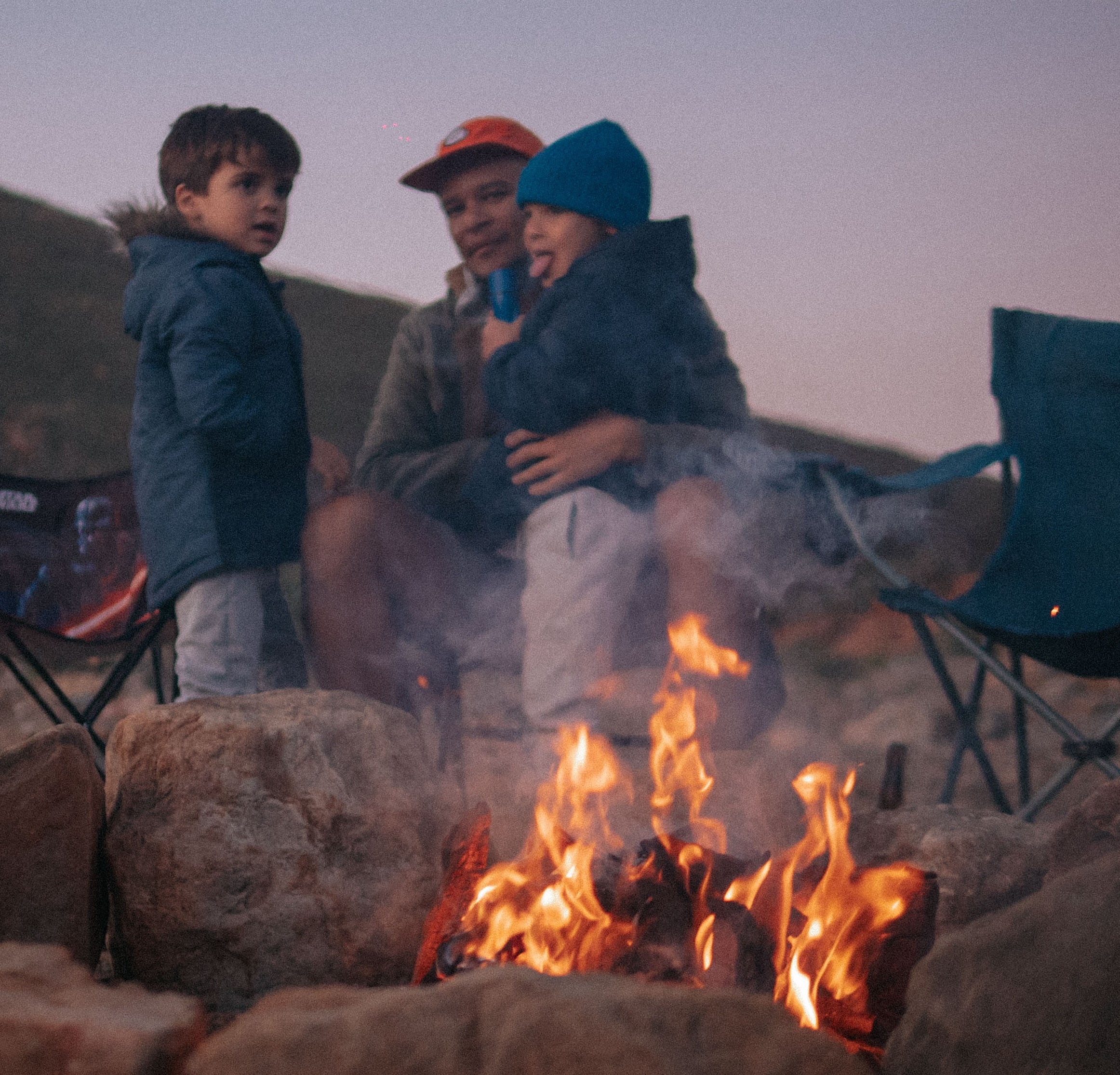 A dad and his two sons and camping chairs behind a fire