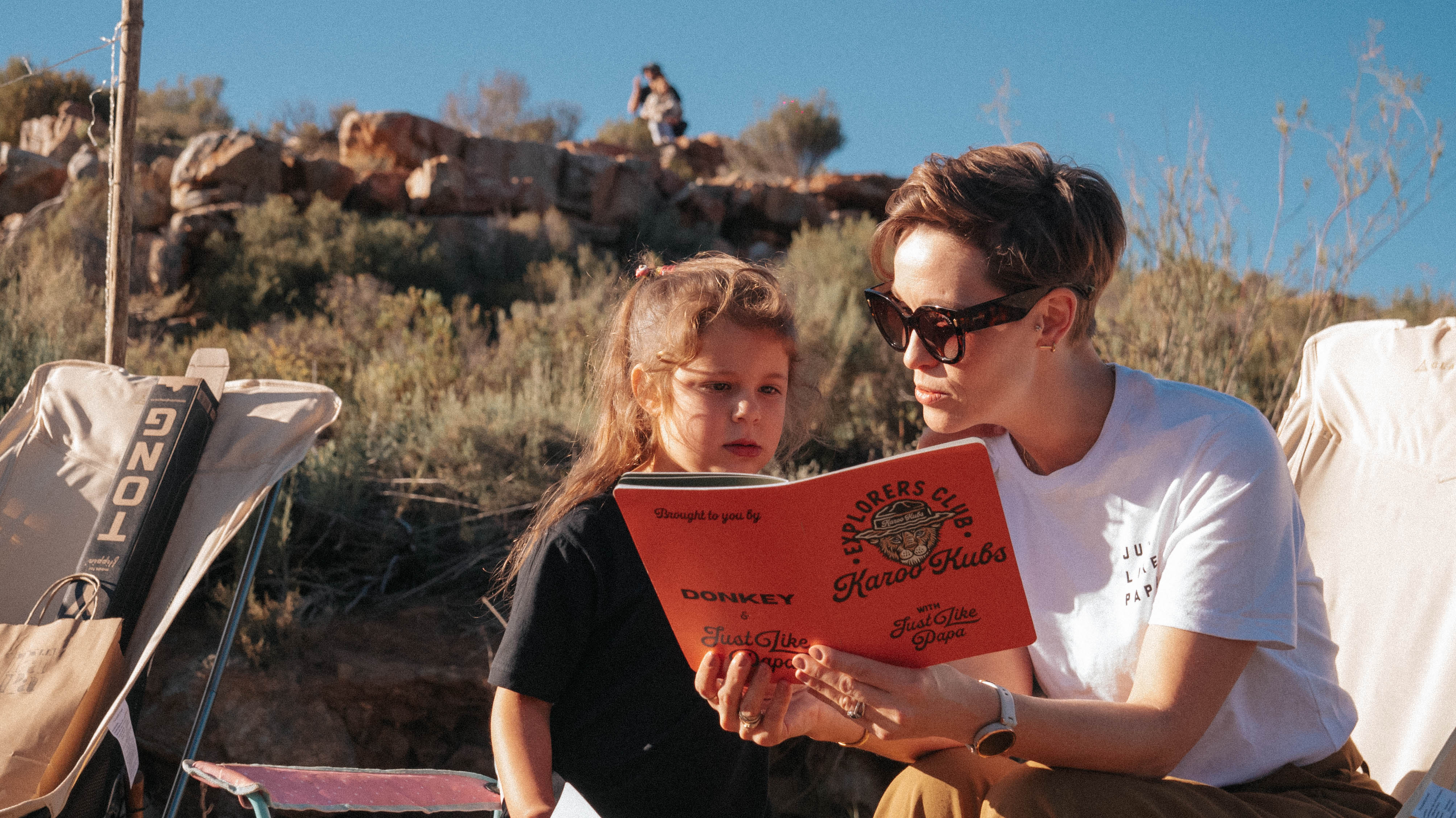 A mom reading through a "Karoo Kubs Explorers Club" book with her daughter in a mountain area